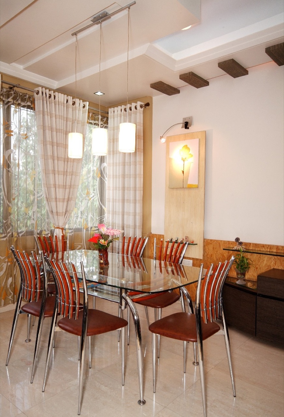 A glass dining table under three pendant lights beside a curtained window